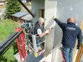 Deux ouvriers en nacelle isolent une façade. L'un perce, l'autre maintient une plaque, luttant contre le déphasage thermique.