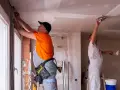Deux ouvriers rénovent un bâtiment, l'un ajuste des câbles près d'une fenêtre, l'autre enduit le plafond. Ces travaux sont essentiels pour les aides aux collectivités locales.