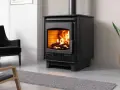 Poêle à bois moderne, flammes vives réchauffant la pièce. Les granulés ne tombent plus, juste du bois qui crépite. Un fauteuil noir et une plante verte complètent la scène.