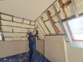 Un ouvrier en combinaison projette de la mousse polyuréthane pour isoler un grenier. L'isolant blanc recouvre les poutres et les murs.