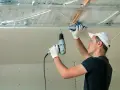 Un ouvrier installe des isolants minces sous un faux plafond en plaques de plâtre, utilisant une perceuse pour fixer les éléments.