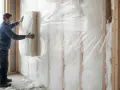 Un ouvrier installe un panneau d'isolant blanc dans un mur intérieur en bois, recouvert d'une bâche plastique transparente.