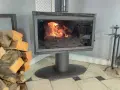 Un poêle à bois avec une vitre propre pour changer une vitre de poele a bois. Des bûches de bois sont empilées à gauche.