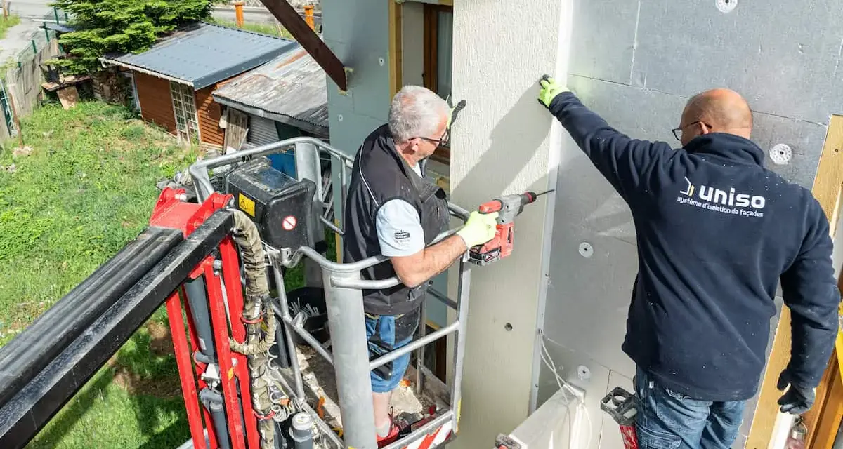 Deux ouvriers en nacelle isolent une façade. L'un perce, l'autre maintient une plaque, luttant contre le déphasage thermique.