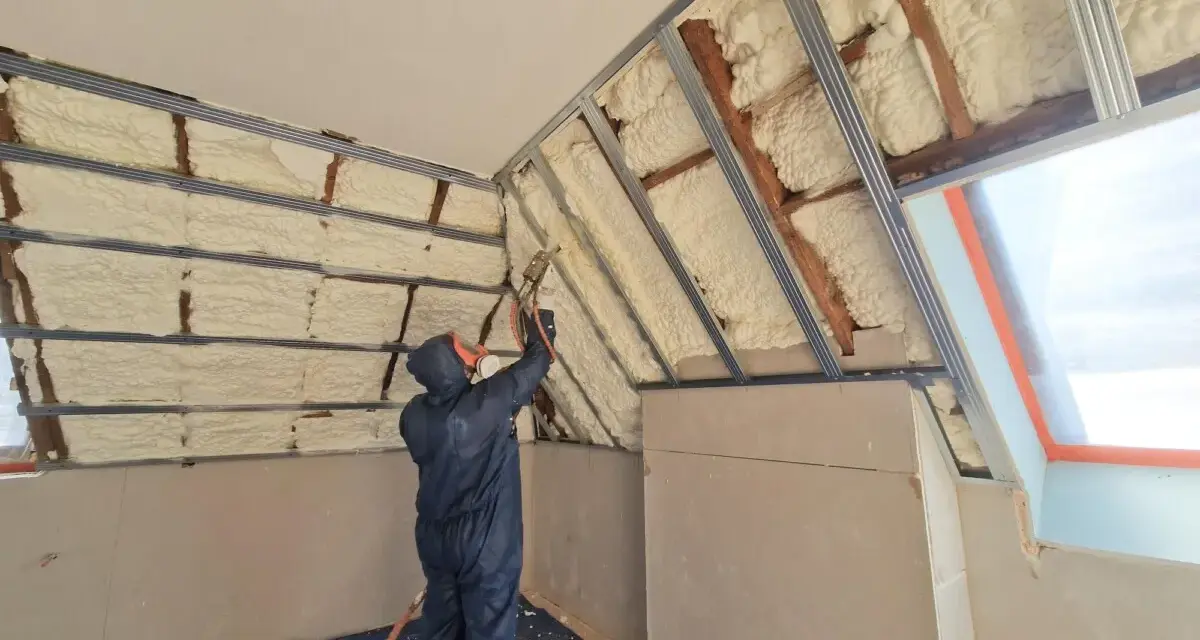 Un ouvrier en combinaison projette de la mousse polyuréthane pour isoler un grenier. L'isolant blanc recouvre les poutres et les murs.