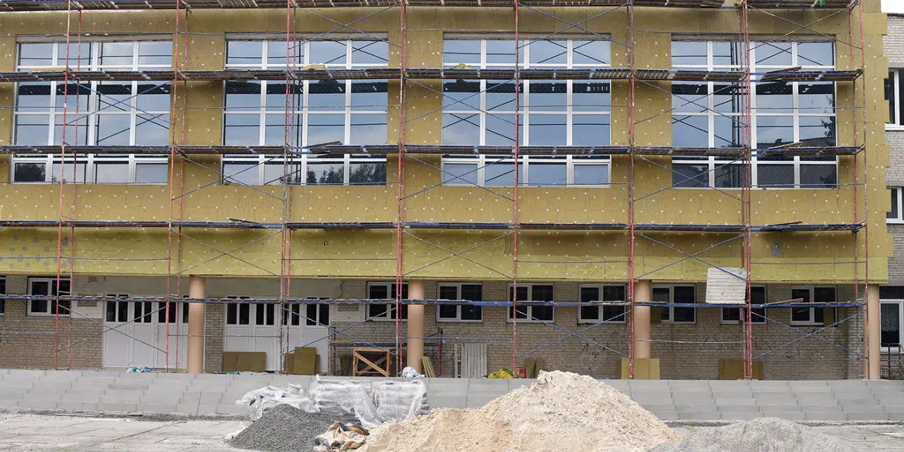 Bâtiment en rénovation énergétique, échafaudages et isolation jaune recouvrent la façade. Des tas de sable et de gravier sont prêts pour les travaux.