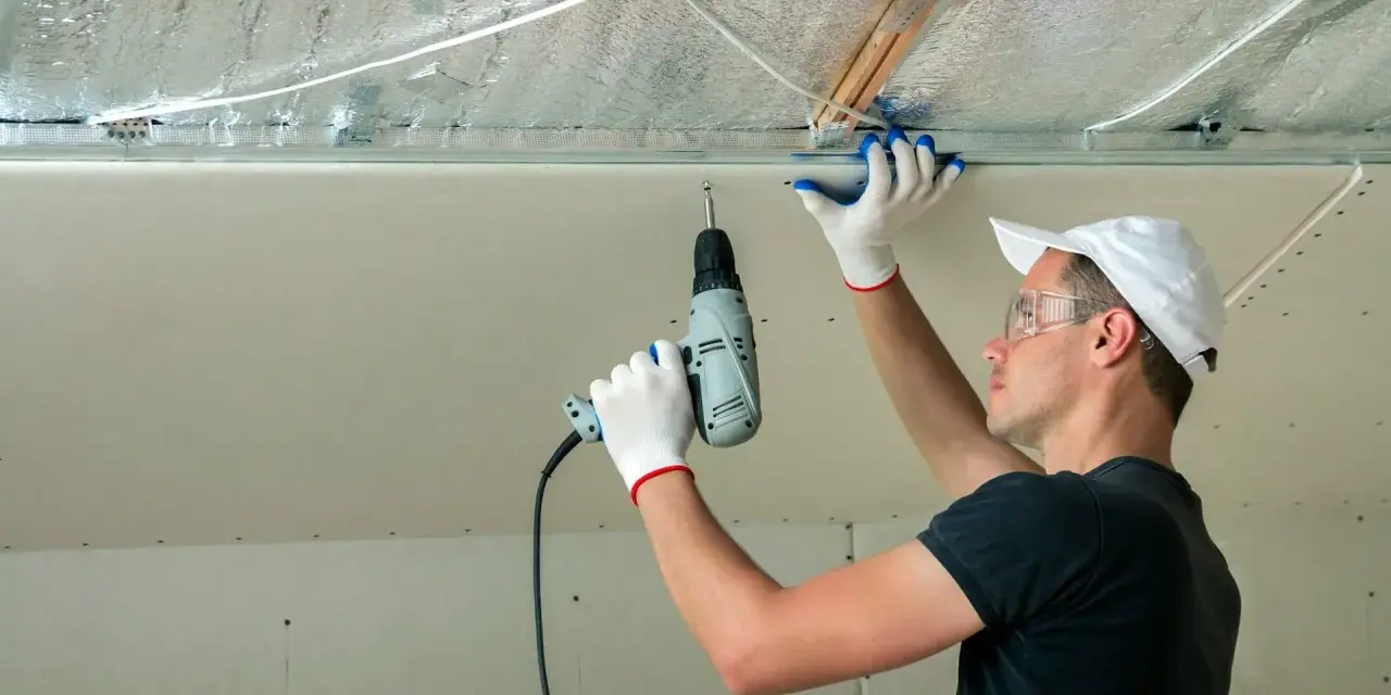 Un ouvrier installe des isolants minces sous un faux plafond en plaques de plâtre, utilisant une perceuse pour fixer les éléments.