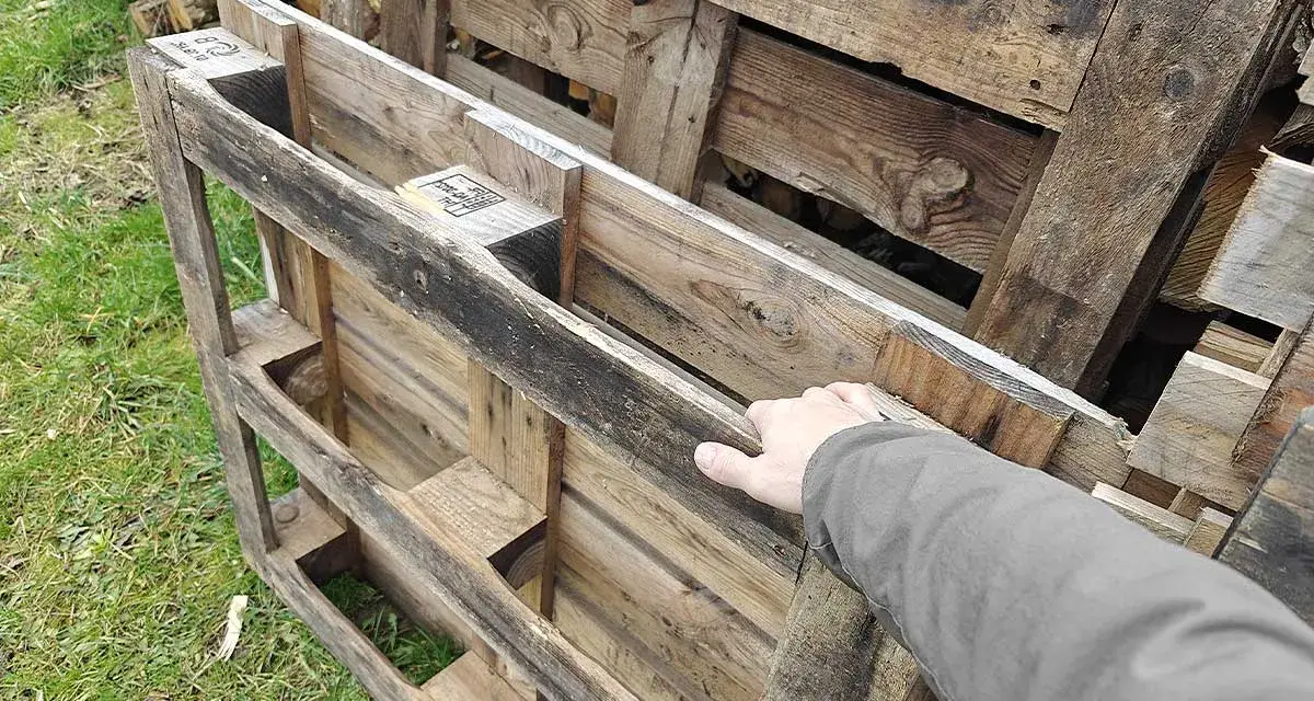 Maintenant, la question se pose : peut-on brûler du bois de palette dans une cheminée ? Une main touche des palettes en bois usagées, empilées sur de l'herbe.