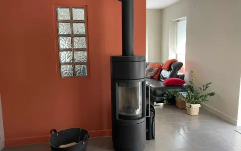 Un poêle moderne noir se dresse devant une peinture murale ocre, à côté d'une fenêtre en verre dépoli. Un canapé en cuir noir et des coussins colorés sont visibles au loin.