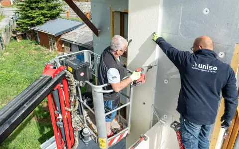Deux ouvriers en nacelle isolent une façade. L'un perce, l'autre maintient une plaque, luttant contre le déphasage thermique.