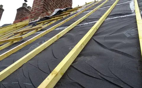 Charpente de toiture avec isolant noir et liteaux en bois, près de cheminées en brique.