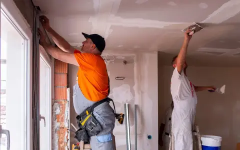 Deux ouvriers rénovent un bâtiment, l'un ajuste des câbles près d'une fenêtre, l'autre enduit le plafond. Ces travaux sont essentiels pour les aides aux collectivités locales.