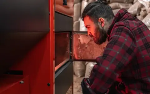 Homme vérifiant un poêle à granulés, peut-être une alarme 6 s'est déclenchée. Sacs de granulés en arrière-plan.