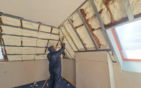 Un ouvrier en combinaison projette de la mousse polyuréthane pour isoler un grenier. L'isolant blanc recouvre les poutres et les murs.