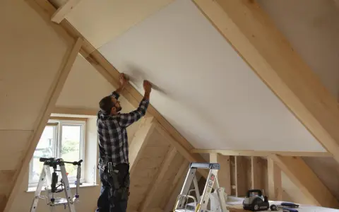 Un ouvrier installe un panneau d'isolant polyuréthane dans un grenier en cours d'aménagement, sous une charpente en bois.