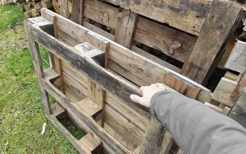 Maintenant, la question se pose : peut-on brûler du bois de palette dans une cheminée ? Une main touche des palettes en bois usagées, empilées sur de l'herbe.