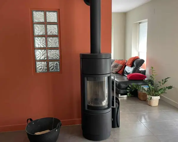 Un poêle moderne noir se dresse devant une peinture murale ocre, à côté d'une fenêtre en verre dépoli. Un canapé en cuir noir et des coussins colorés sont visibles au loin.
