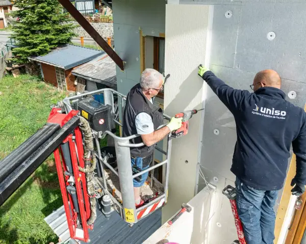 Deux ouvriers en nacelle isolent une façade. L'un perce, l'autre maintient une plaque, luttant contre le déphasage thermique.