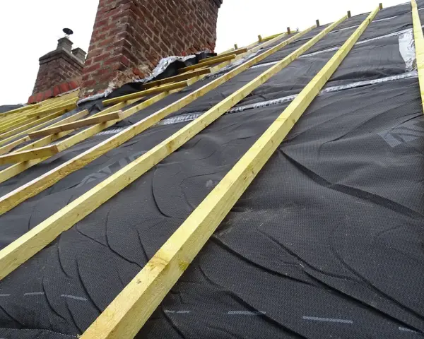 Charpente de toiture avec isolant noir et liteaux en bois, près de cheminées en brique.