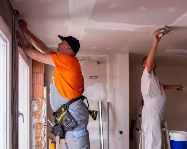 Deux ouvriers rénovent un bâtiment, l'un ajuste des câbles près d'une fenêtre, l'autre enduit le plafond. Ces travaux sont essentiels pour les aides aux collectivités locales.