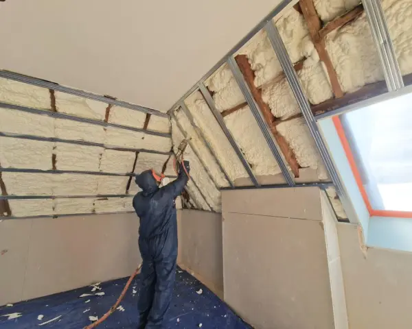 Un ouvrier en combinaison projette de la mousse polyuréthane pour isoler un grenier. L'isolant blanc recouvre les poutres et les murs.