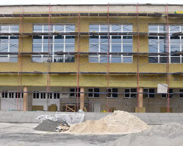 Bâtiment en rénovation énergétique, échafaudages et isolation jaune recouvrent la façade. Des tas de sable et de gravier sont prêts pour les travaux.