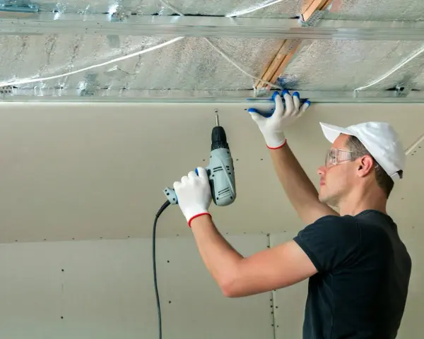 Un ouvrier installe des isolants minces sous un faux plafond en plaques de plâtre, utilisant une perceuse pour fixer les éléments.