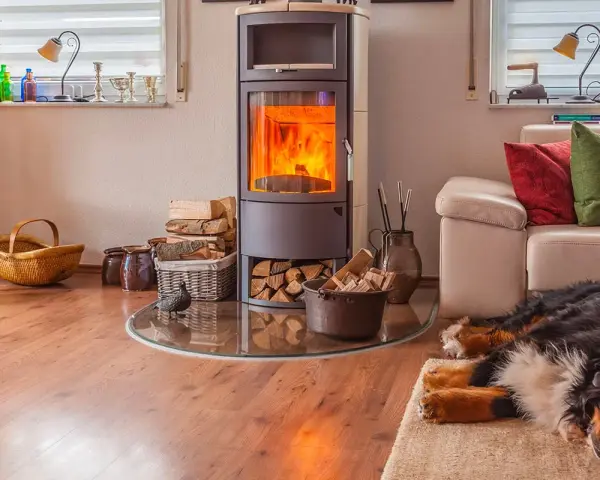 Un poêle bouilleur mixte crépite, réchauffant le salon où un chien dort paisiblement sur un tapis. Bois et paniers complètent ce tableau cosy.