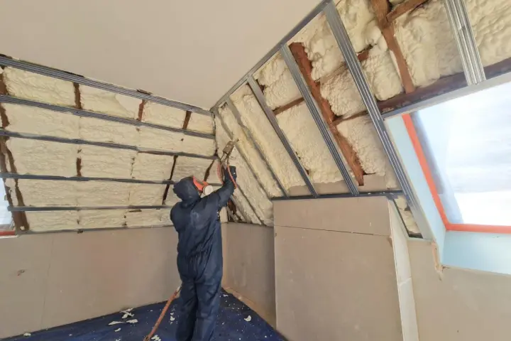 Un ouvrier en combinaison projette de la mousse polyuréthane pour isoler un grenier. L'isolant blanc recouvre les poutres et les murs.