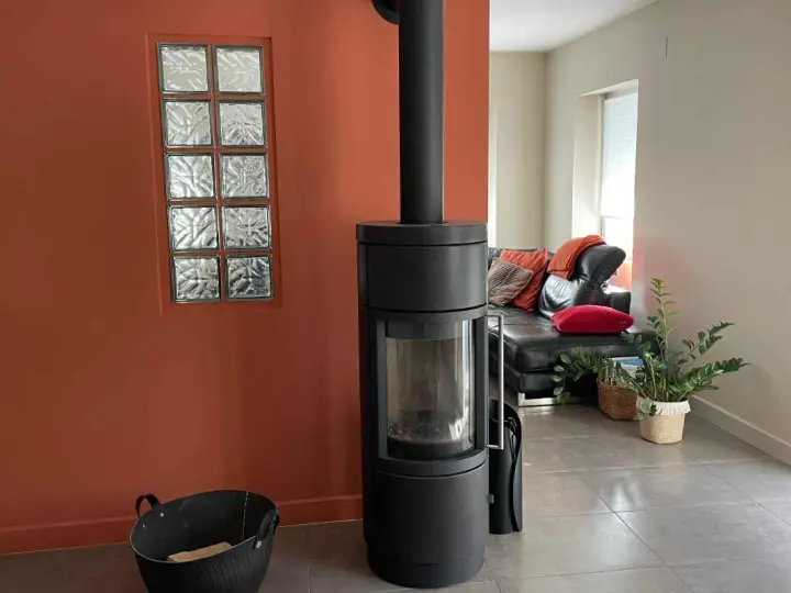 Un poêle moderne noir se dresse devant une peinture murale ocre, à côté d'une fenêtre en verre dépoli. Un canapé en cuir noir et des coussins colorés sont visibles au loin.