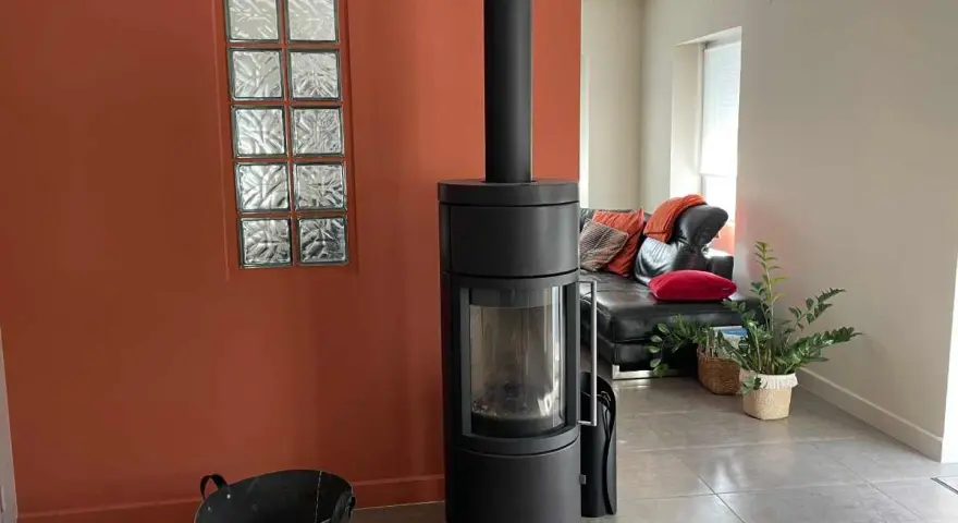 Un poêle moderne noir se dresse devant une peinture murale ocre, à côté d'une fenêtre en verre dépoli. Un canapé en cuir noir et des coussins colorés sont visibles au loin.
