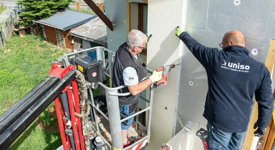 Deux ouvriers en nacelle isolent une façade. L'un perce, l'autre maintient une plaque, luttant contre le déphasage thermique.
