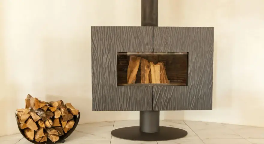 Un poêle moderne avec une cheminée vitrée et du bois de chauffage dans un panier. La **déco derrière un poêle** est sobre, mettant en valeur l'âtre.