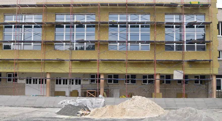 Bâtiment en rénovation énergétique, échafaudages et isolation jaune recouvrent la façade. Des tas de sable et de gravier sont prêts pour les travaux.