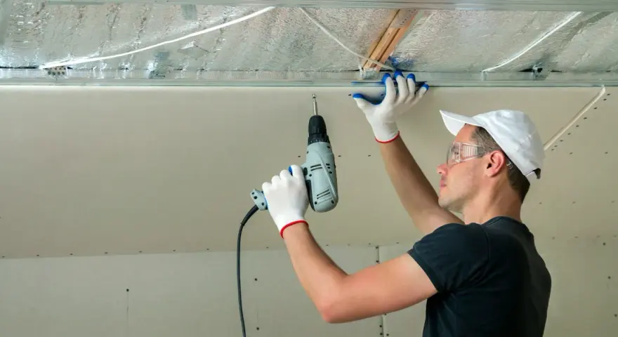 Un ouvrier installe des isolants minces sous un faux plafond en plaques de plâtre, utilisant une perceuse pour fixer les éléments.