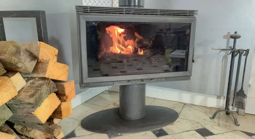 Un poêle à bois avec une vitre propre pour changer une vitre de poele a bois. Des bûches de bois sont empilées à gauche.