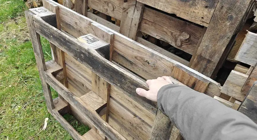 Maintenant, la question se pose : peut-on brûler du bois de palette dans une cheminée ? Une main touche des palettes en bois usagées, empilées sur de l'herbe.
