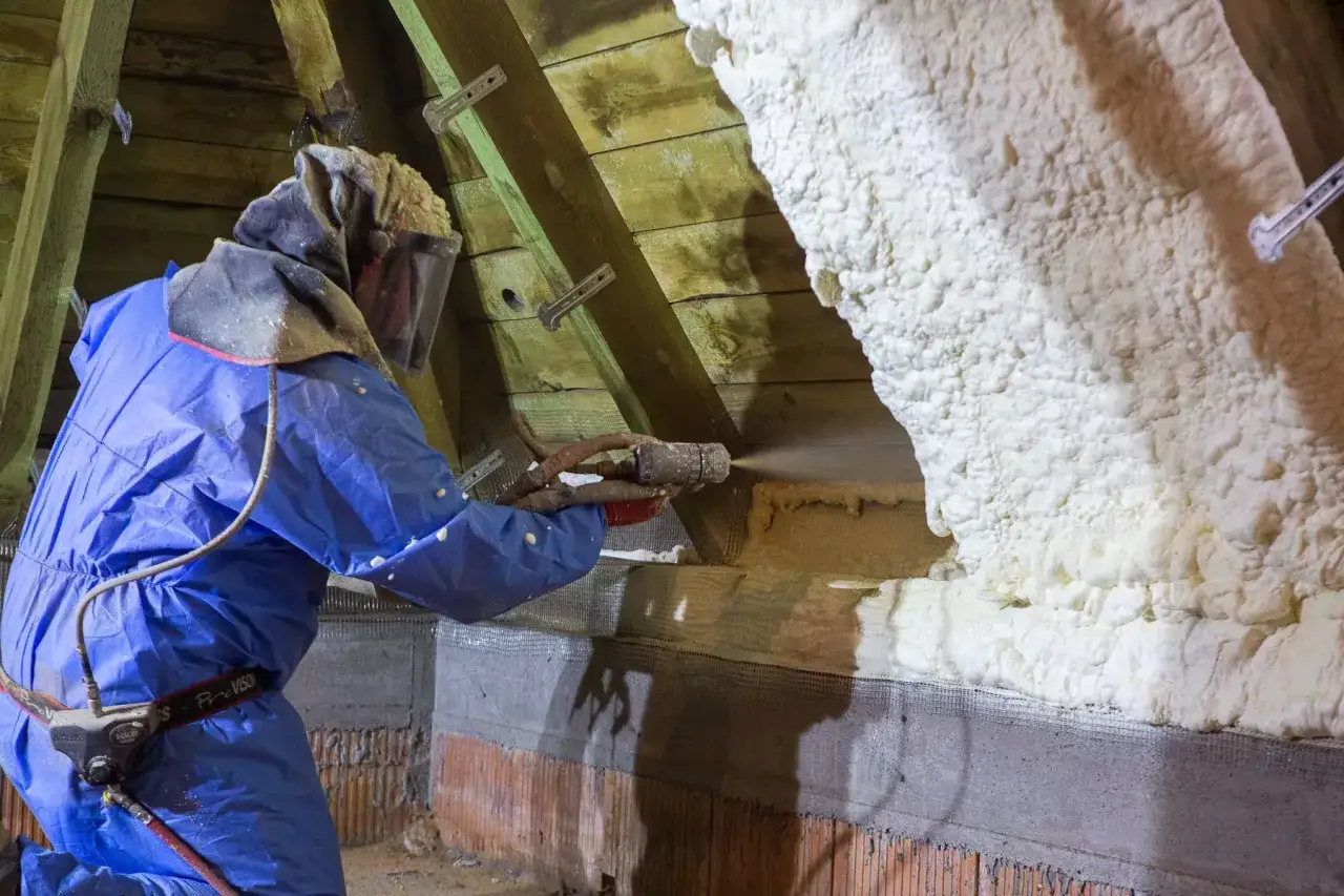 Un ouvrier en combinaison bleue projette de la mousse polyuréthane pour isoler un grenier.