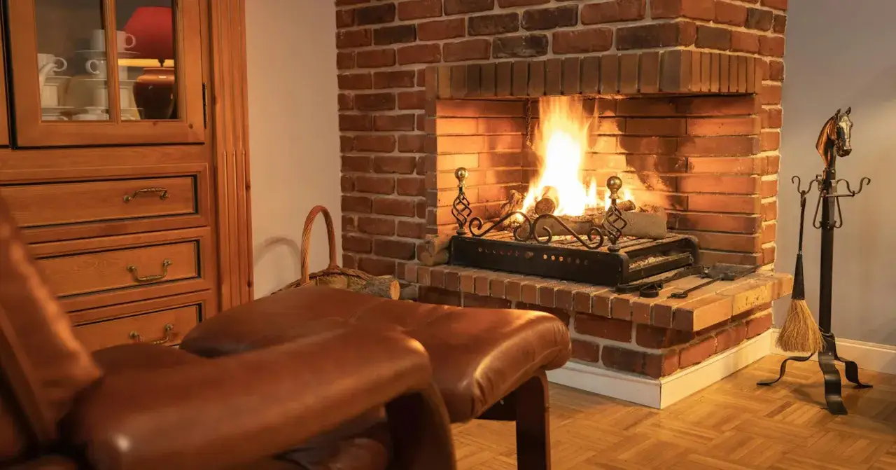 Un poêle moderne est installé dans une ancienne cheminée en briques, créant une ambiance chaleureuse. Un fauteuil en cuir marron et une armoire en bois complètent la scène.