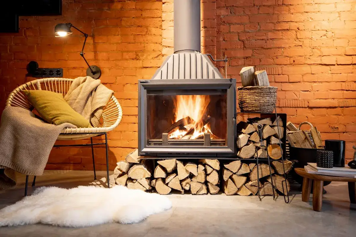 Un po&ecirc;le moderne remplace une ancienne chemin&eacute;e, avec du bois empil&eacute; et un fauteuil confortable pr&egrave;s du feu.